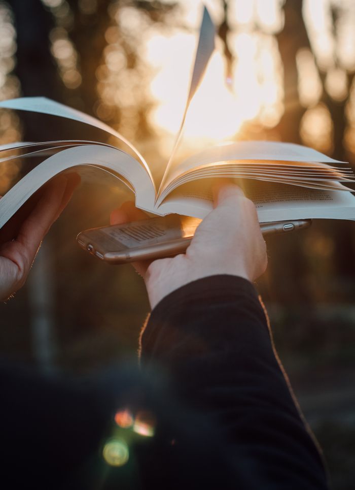 about-img Close-up of hands holding a book and smartphone with sunlight filtering through the trees.