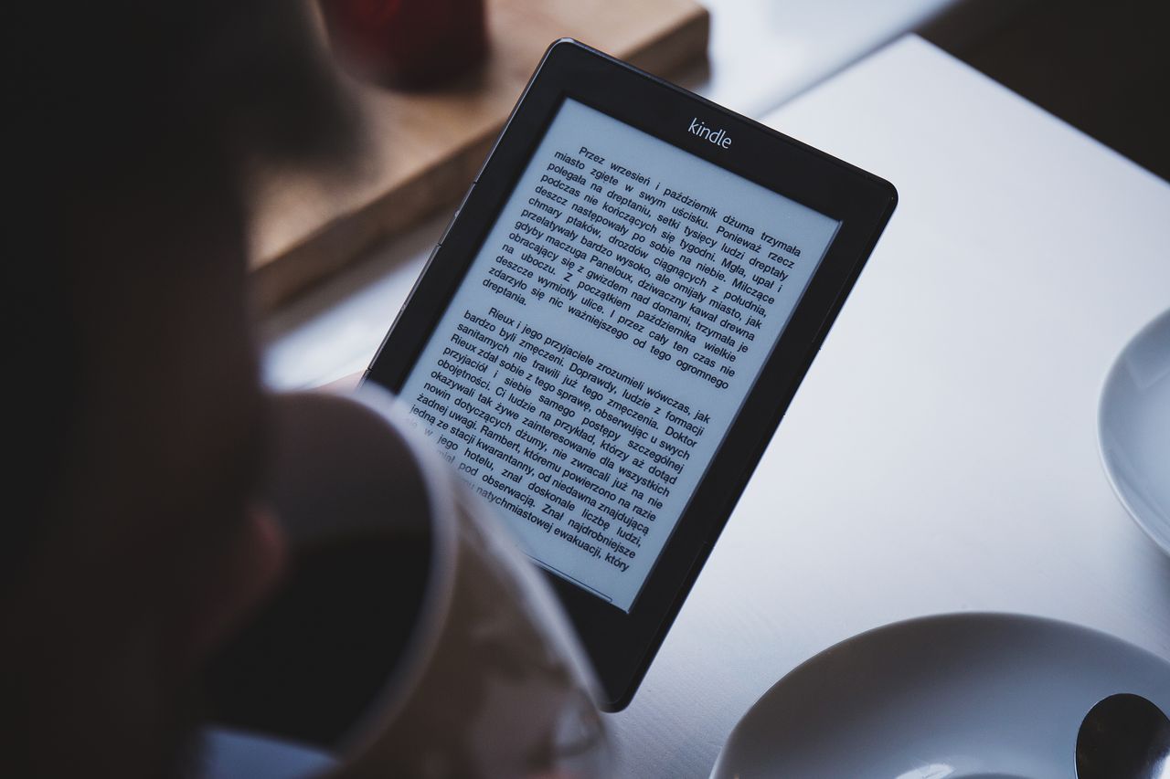 social-feed-img-01 A person reads on a Kindle e-reader while holding a cup of coffee, indoors.